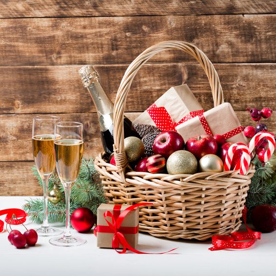 Two glasses of champagne with christmas basket with bottle, gifts with red satin ribbon, candy canes, pine cones, golden garlands on dark brown wooden background, new year celebration concept