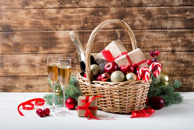 Two glasses of champagne with christmas basket with bottle, gifts with red satin ribbon, candy canes, pine cones, golden garlands on dark brown wooden background, new year celebration concept